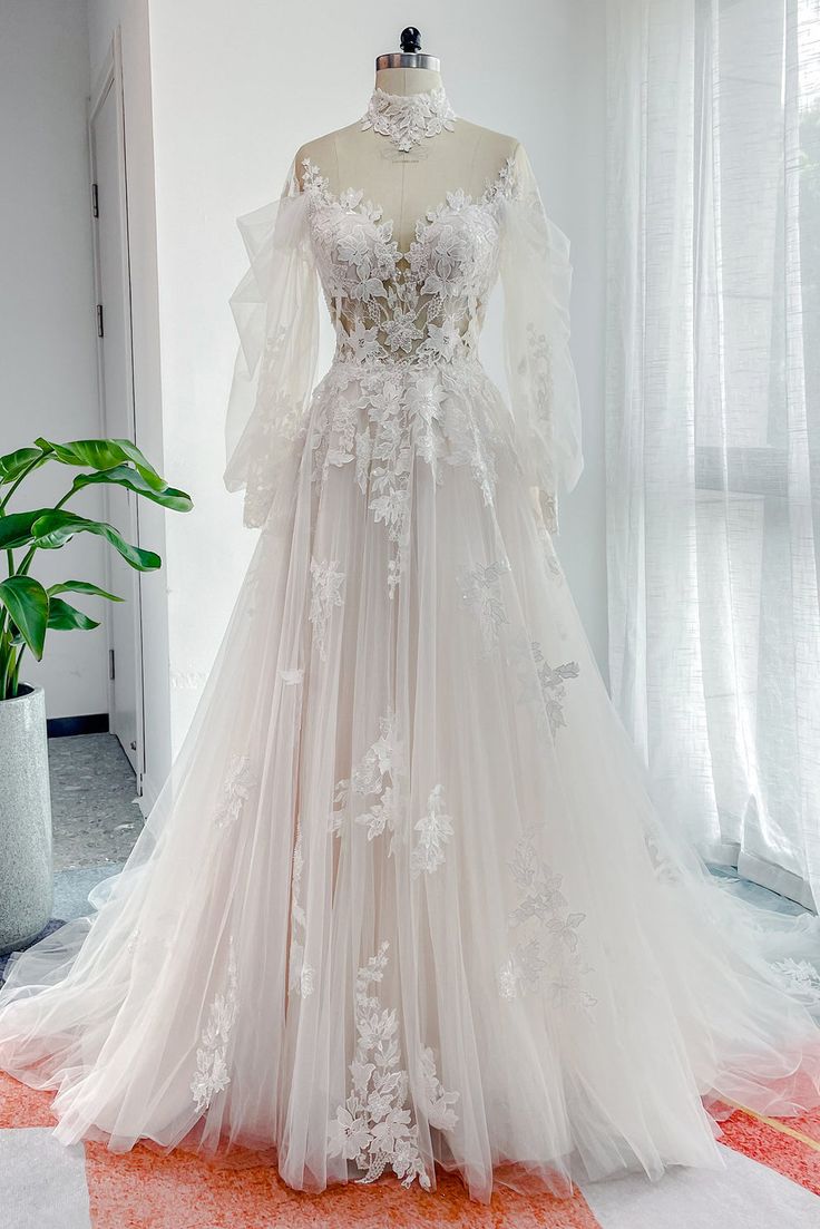 a white wedding dress on display in front of a large potted plant and window