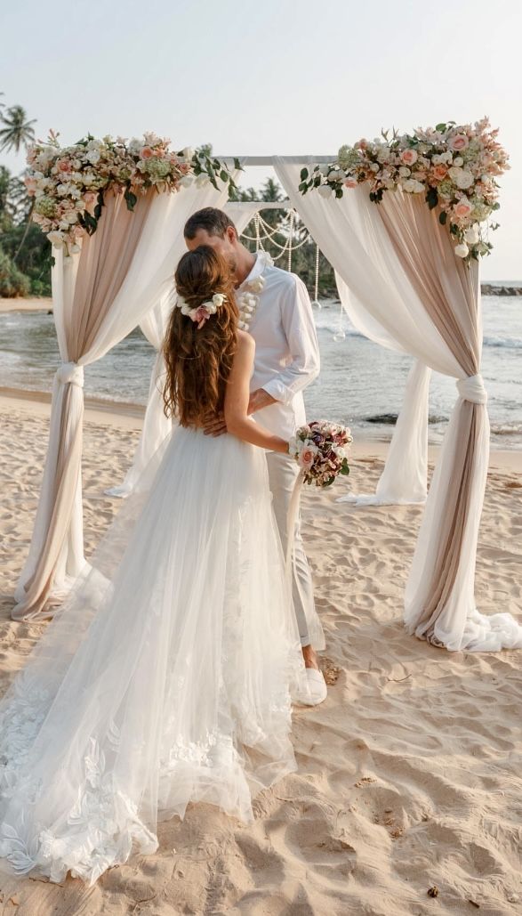 a bride and groom kissing on the beach under an arch decorated with flowers, greenery and ribbons