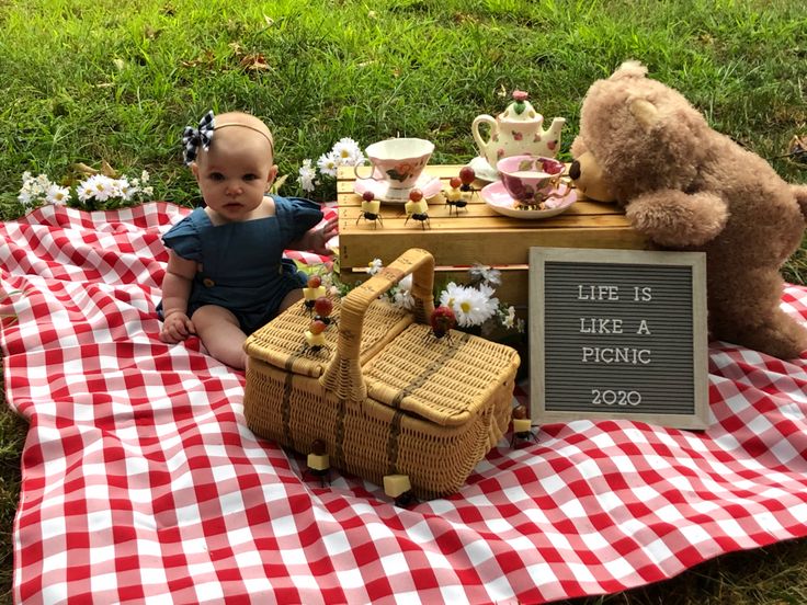 a baby sitting on a picnic blanket next to a teddy bear and some tea set