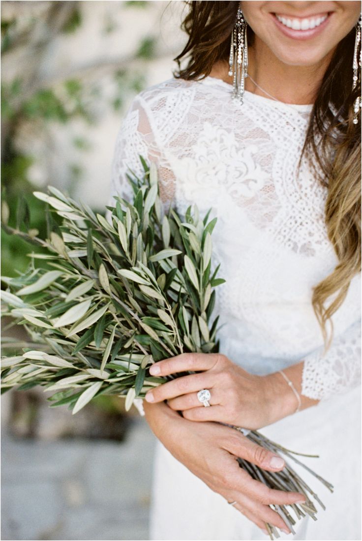 a woman in a white dress holding a bouquet of greenery and smiling at the camera