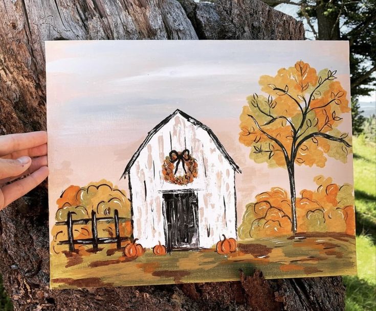 a person holding up a painting of a white barn with autumn trees in the background