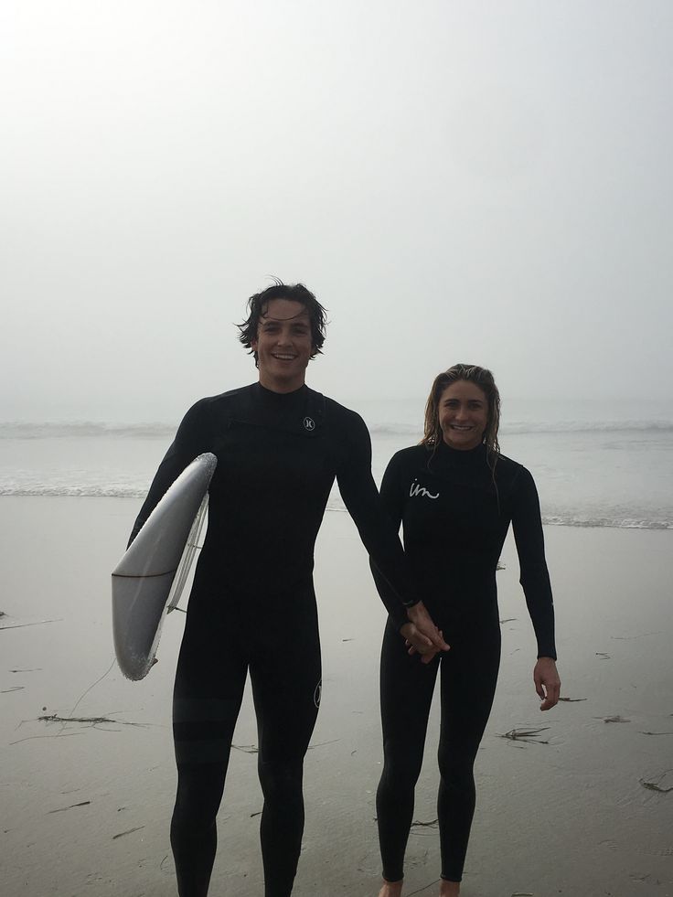 two people in wetsuits standing on the beach holding surfboards and smiling at the camera