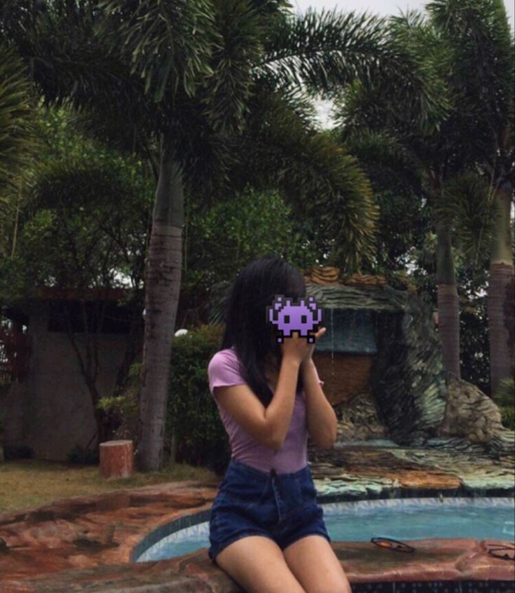 a woman sitting on the edge of a swimming pool talking on her cell phone while holding an object in front of her face