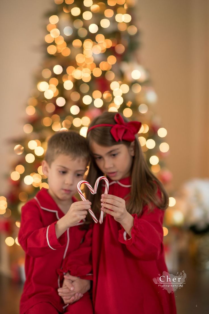 two children in front of a christmas tree holding up heart shaped candy canes while looking at the camera