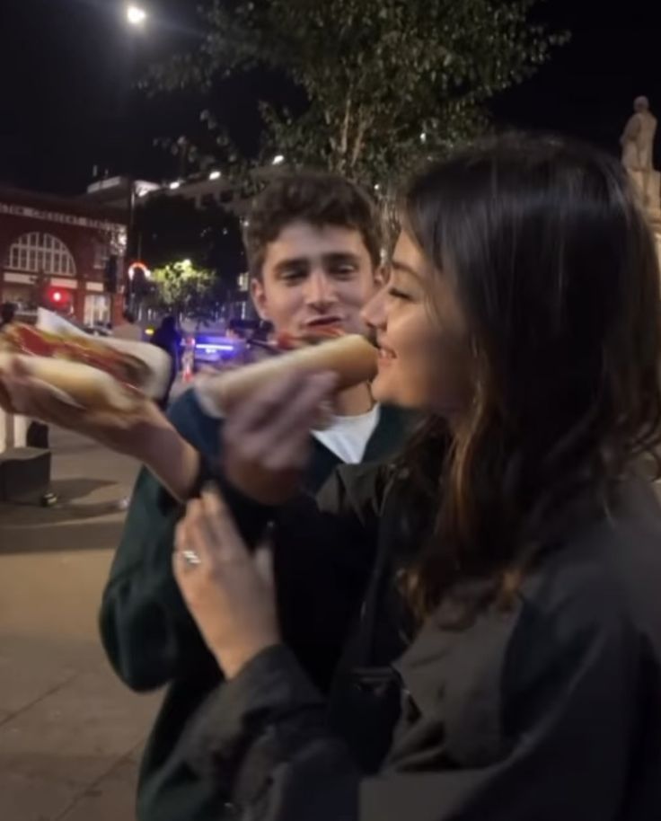 two people eating hotdogs on a city street at night