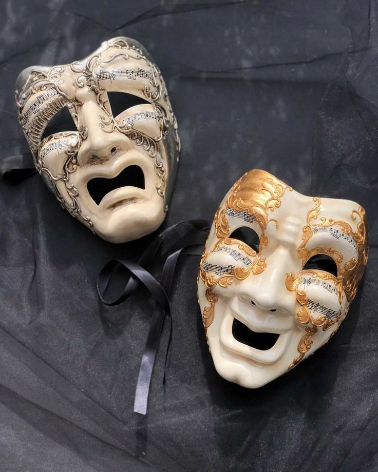 two masquerade masks are sitting on a black cloth, one is white and the other is gold