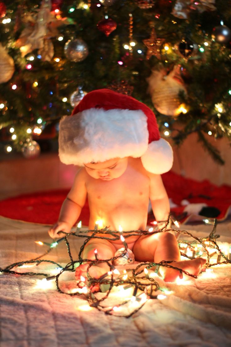 a baby wearing a santa hat sitting in front of a christmas tree with lights on it