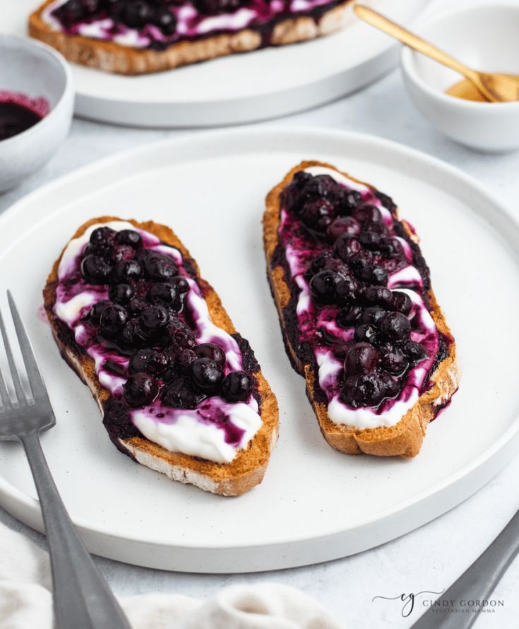 two pieces of bread with blueberries and cream on them sitting on a white plate