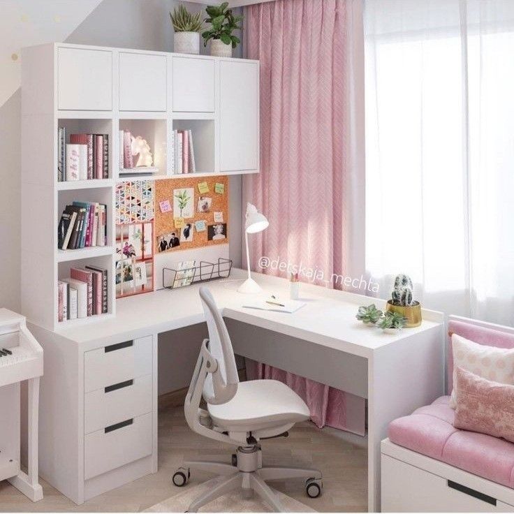 a white desk sitting under a window next to a pink chair in front of a book shelf