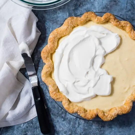 a pie with whipped cream on top sitting next to a knife and bowl of sauce