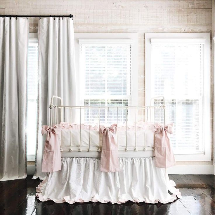 a white crib with pink ruffled bedding in front of two large windows