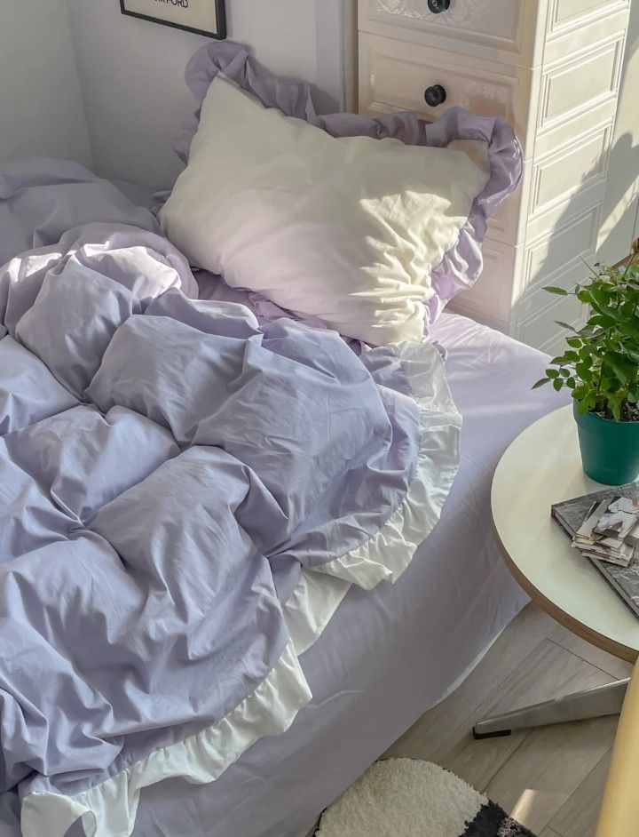 an unmade bed with purple sheets and white pillows in a small room next to a plant