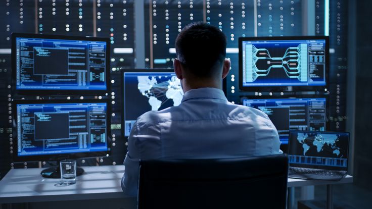 a man sitting at a desk in front of two computer monitors with multiple screens on them