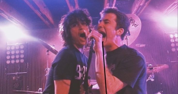 two young men singing into microphones in front of purple lights at a concert venue