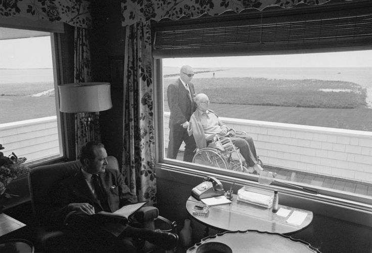 an elderly man sitting in a chair looking out the window at another person standing next to him