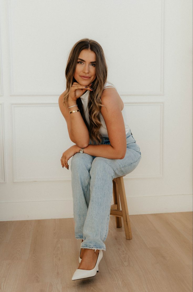 a woman sitting on top of a wooden chair next to a white wall and holding her hand under her chin