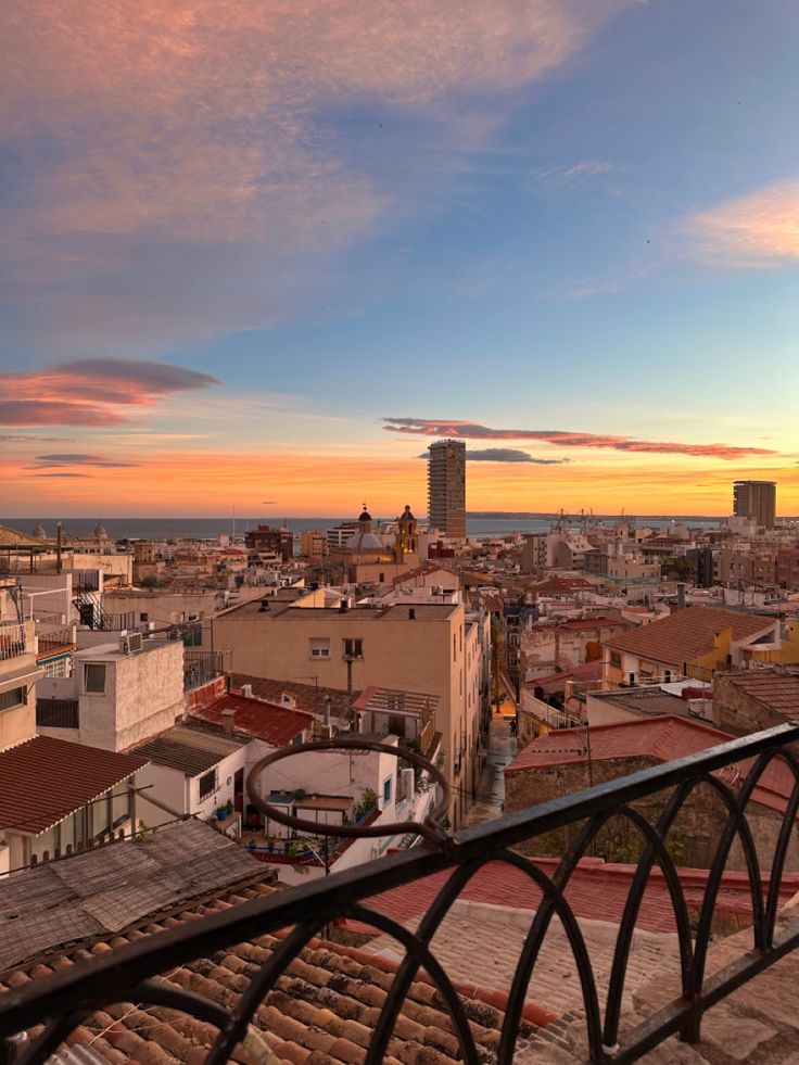 the sun is setting over an urban area with buildings and water in the distance, as seen from a balcony