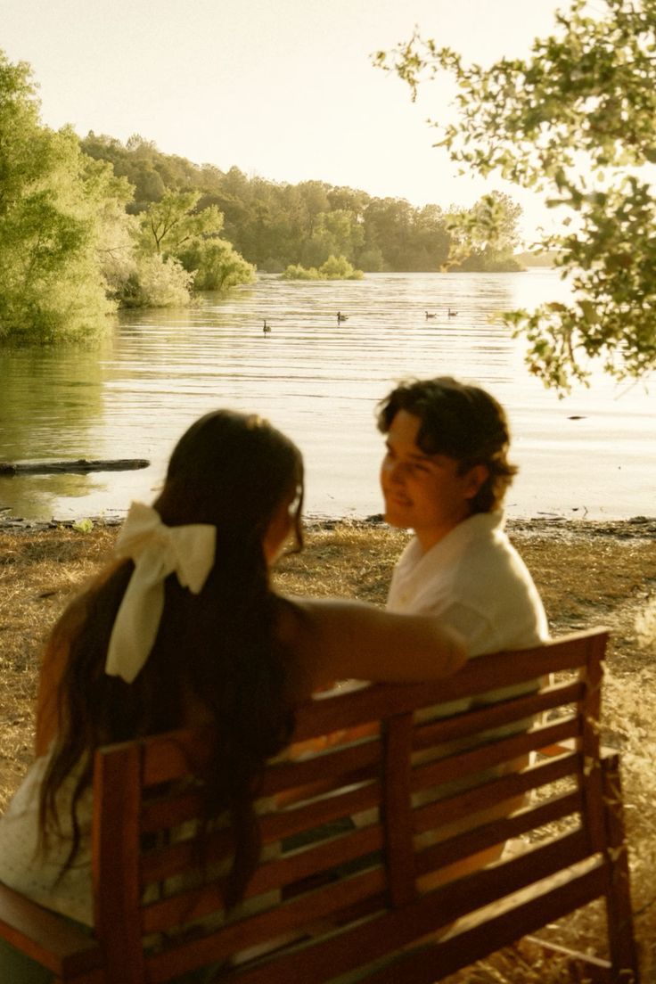 two people are sitting on a bench by the water looking at each other and smiling