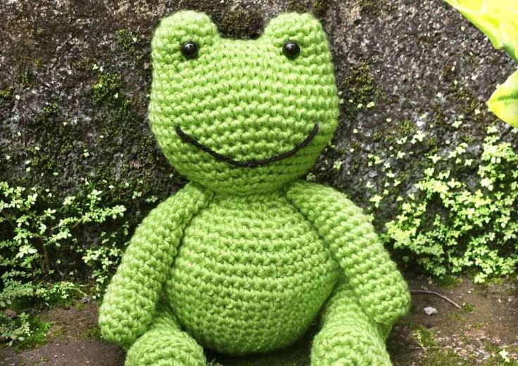 a crocheted frog sitting on the ground next to a wall with moss growing around it