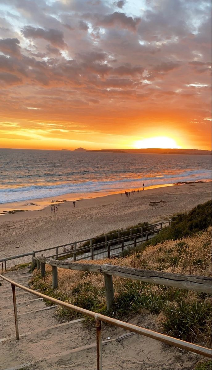 the sun is setting over the beach and people are walking on the path to the water