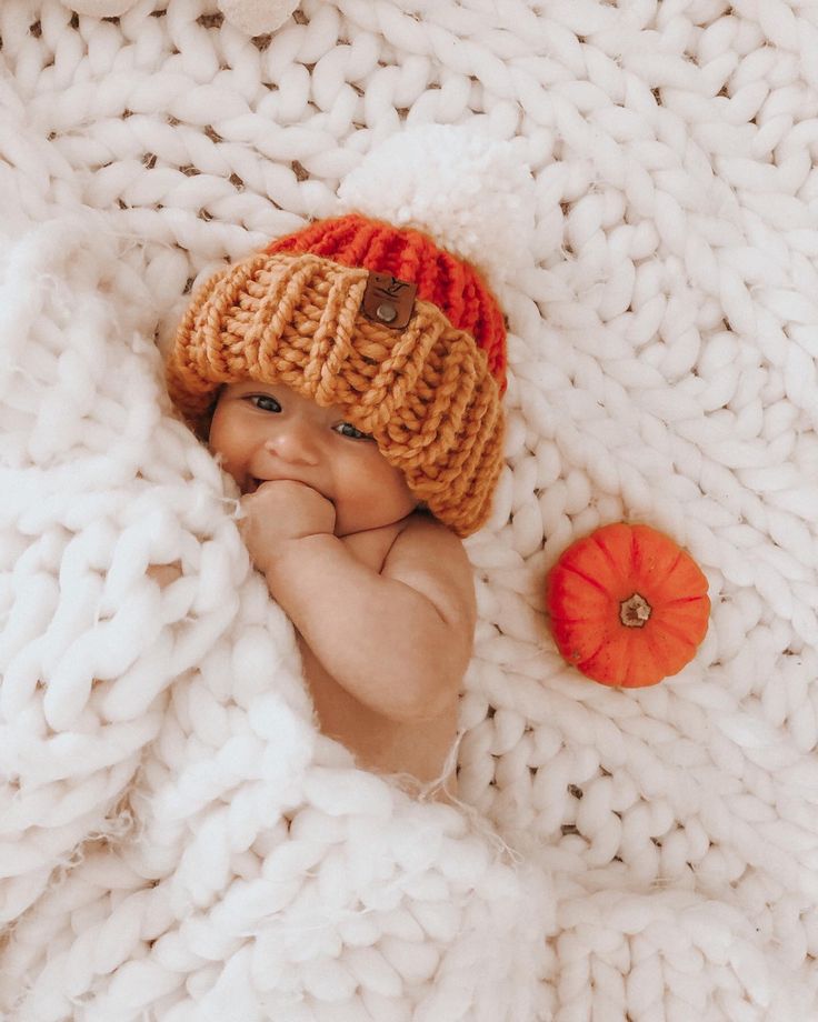 a baby wearing a knitted hat laying on top of a blanket next to an orange pumpkin