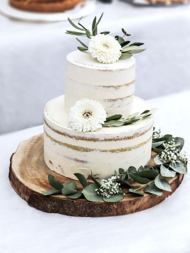 a white wedding cake with flowers and greenery sits on a wooden slice in front of other desserts