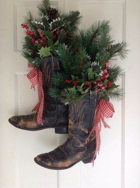 two cowboy boots with christmas decorations hanging from the side of a door, decorated with holly and pine cones