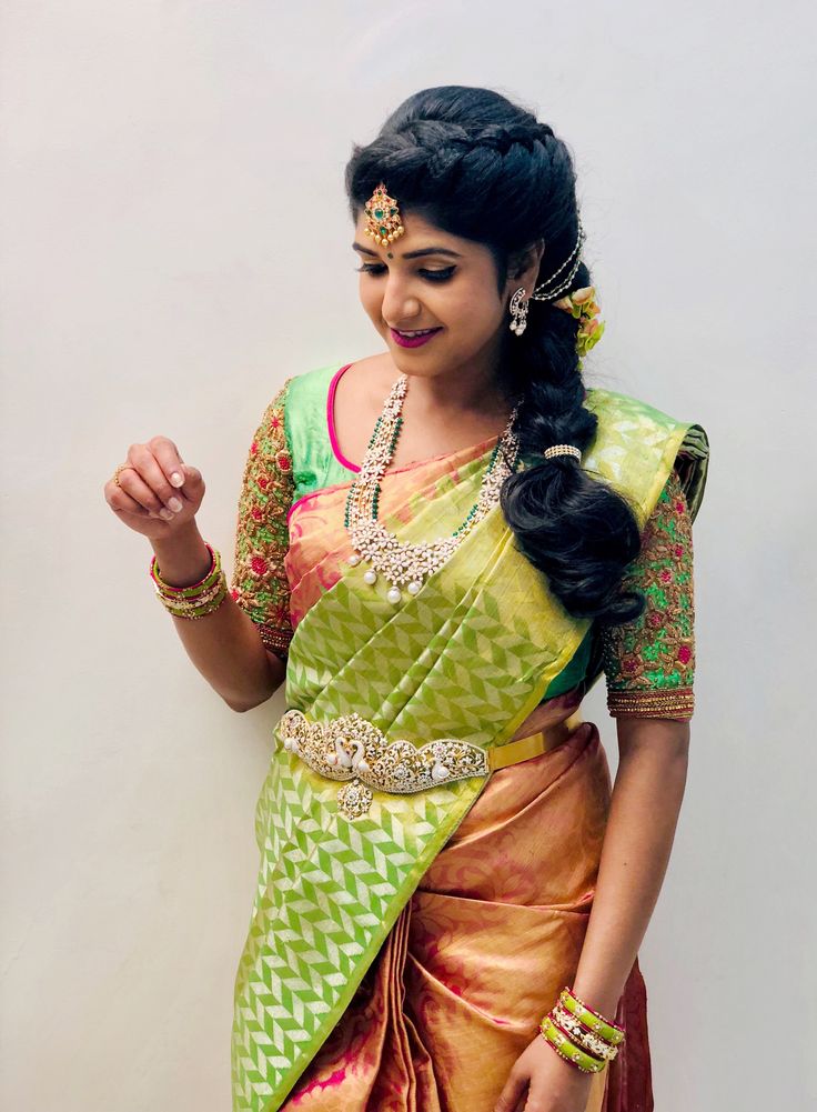 a woman in a green and yellow sari with gold jewelry on her neck, standing next to a white wall