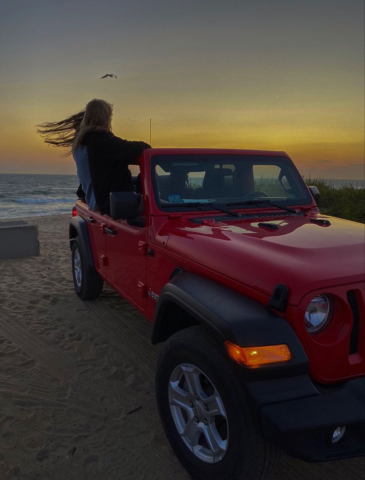 a woman sitting in the back of a red jeep on top of a sandy beach