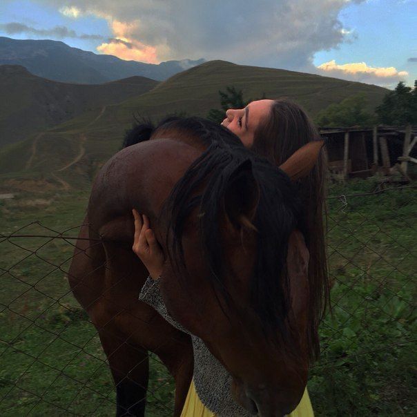 a woman standing next to a brown horse on top of a lush green field under a cloudy sky