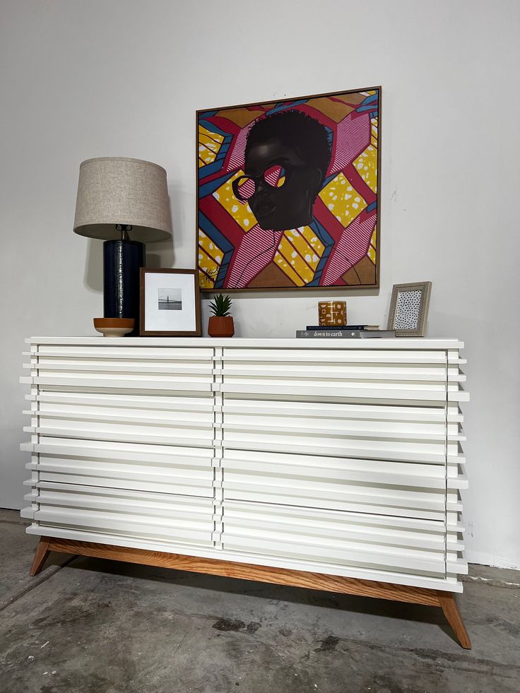 a white cabinet sitting next to a lamp on top of a wooden table in front of a painting