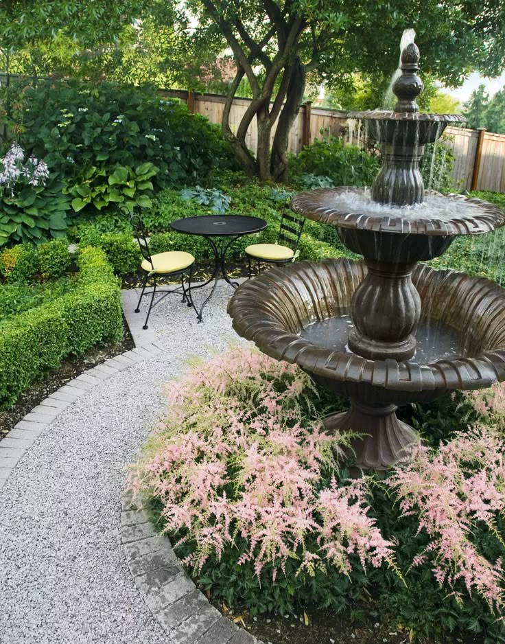 a fountain in the middle of a garden with tables and chairs around it, surrounded by greenery