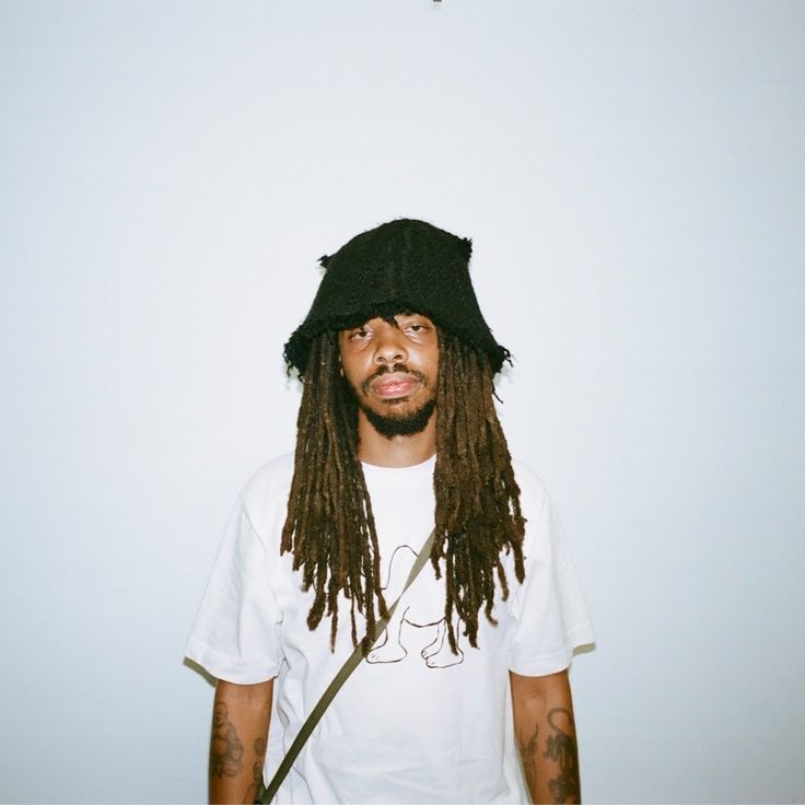 a man with dreadlocks and a hat on his head is standing in front of a white wall