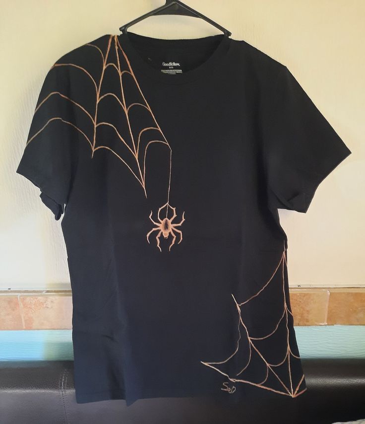 a black t - shirt with a spider on it hanging from a hanger in front of a wall