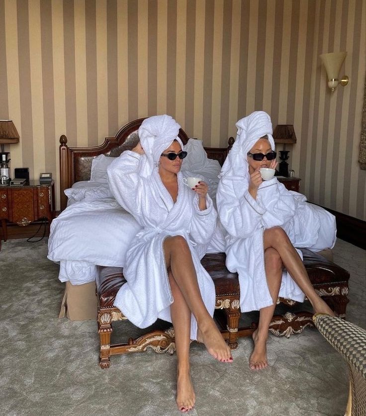 two women in robes are sitting on a bed and one is holding a coffee cup