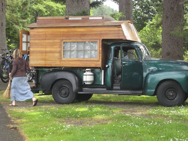 a woman walking past an old green truck with a wooden cabin on the back and windows