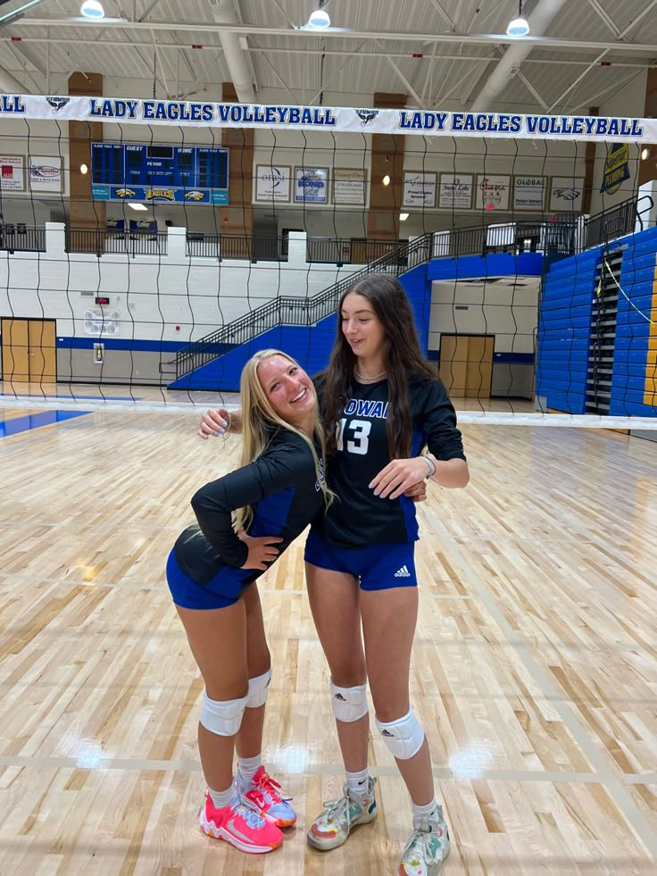 two girls in volleyball uniforms standing on a court with their arms around each other and smiling