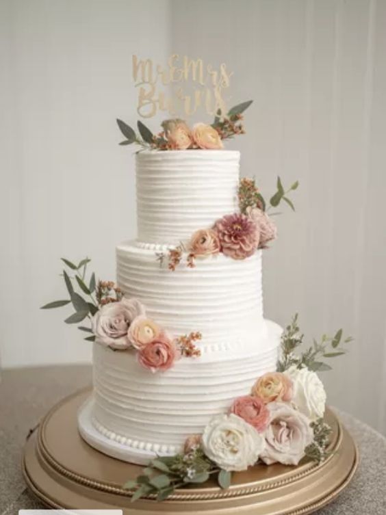 a white wedding cake with flowers and greenery on the top is sitting on a gold platter