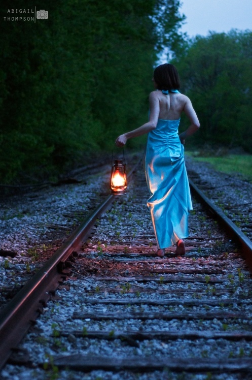 a woman in a blue dress is walking on train tracks with a lantern at night