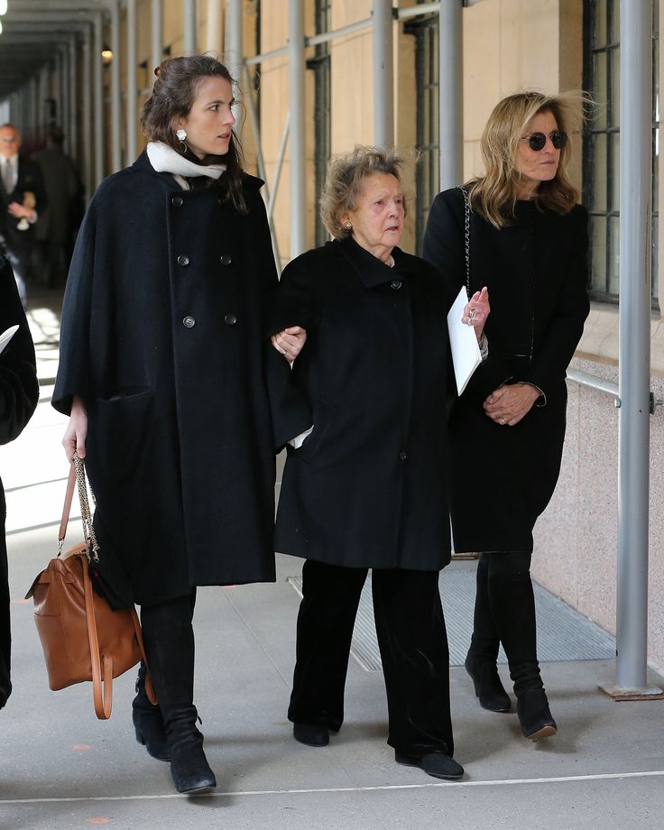 three women are walking down the sidewalk in black coats and holding purses while one woman is on her cell phone