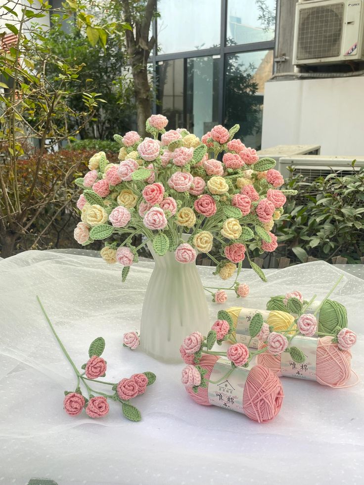 a white vase filled with pink and yellow flowers next to crochet spools