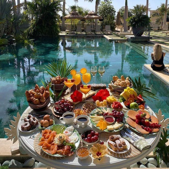 a table filled with lots of food next to a pool in the middle of palm trees