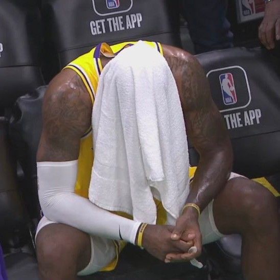 a basketball player sitting on the sidelines with his head in his hands while holding a towel