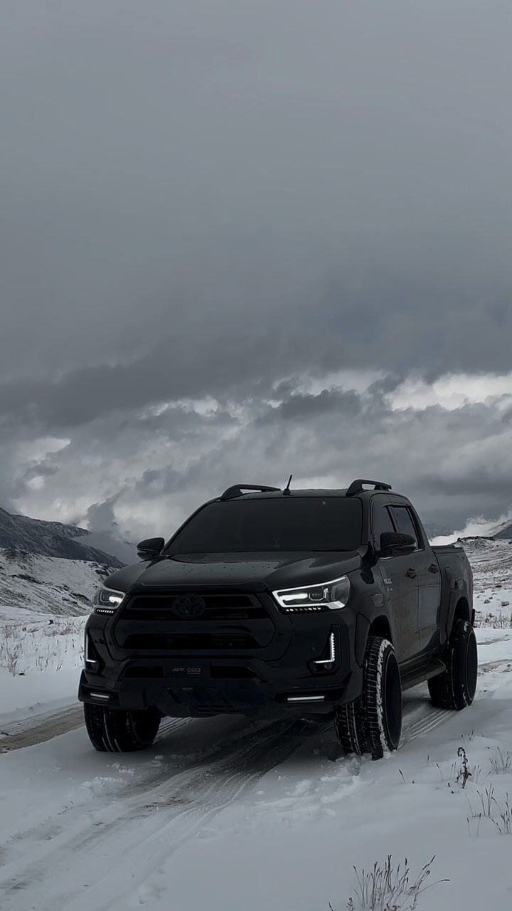 a black truck driving down a snow covered road in the middle of winter with dark clouds overhead