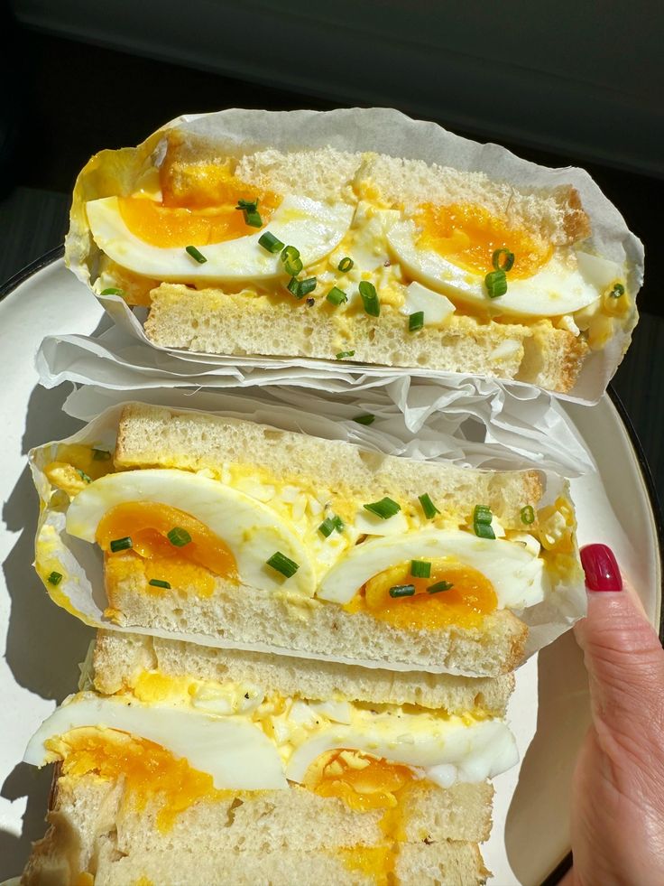 two sandwiches with eggs and cheese are on a white plate, being held by a woman's hand