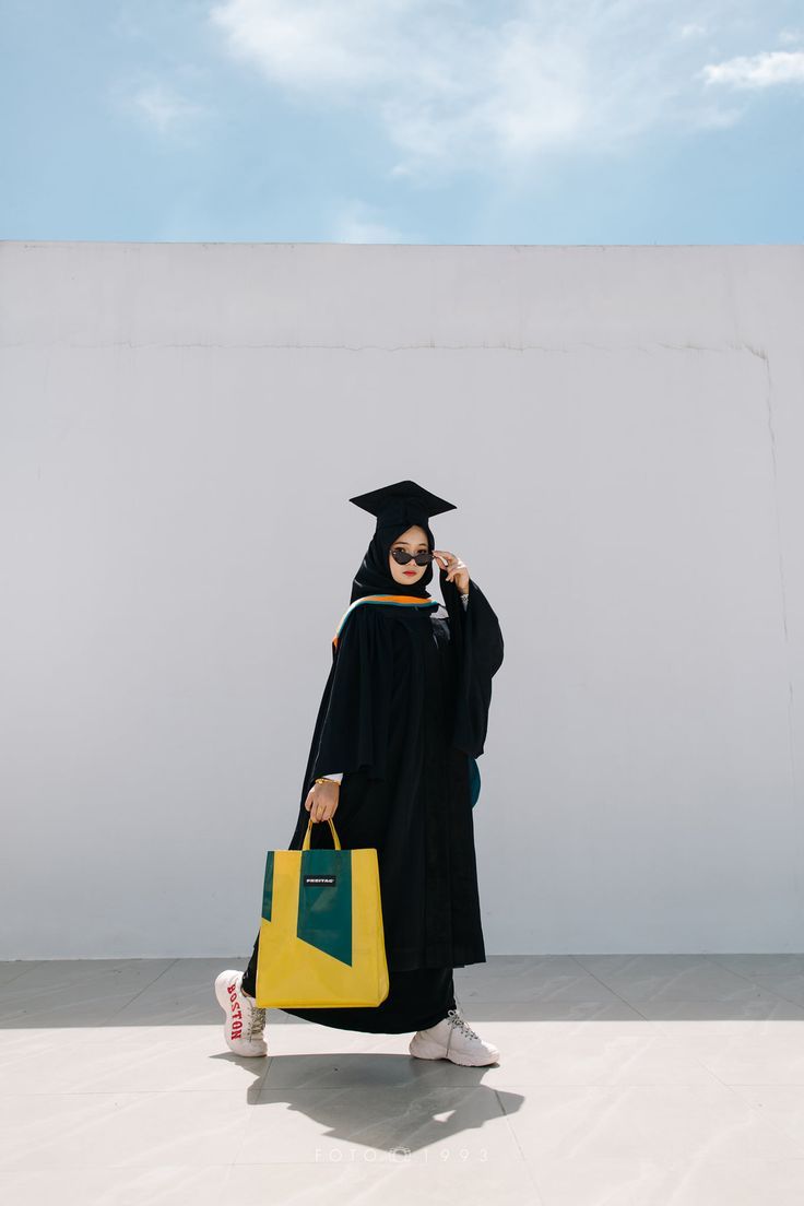 a woman wearing a graduation gown and holding a yellow bag while standing in front of a white wall