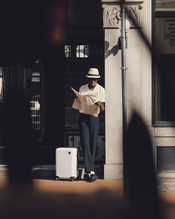 a man standing on the sidewalk reading a newspaper next to a white suit case and suitcase