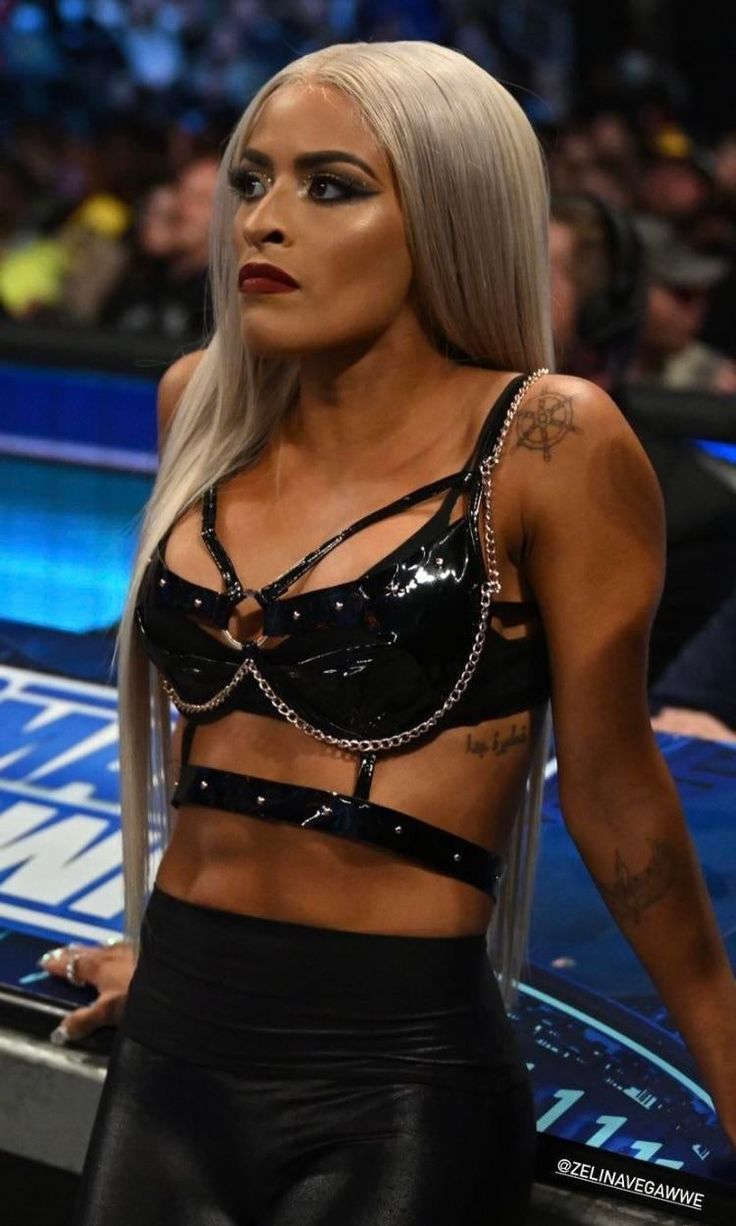 a woman with blonde hair and tattoos standing in front of a wrestling ring wearing black leather pants