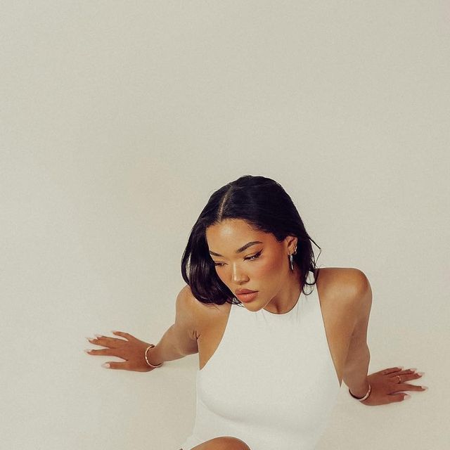 a woman in a white bodysuit sitting on the floor with her arms outstretched and legs crossed