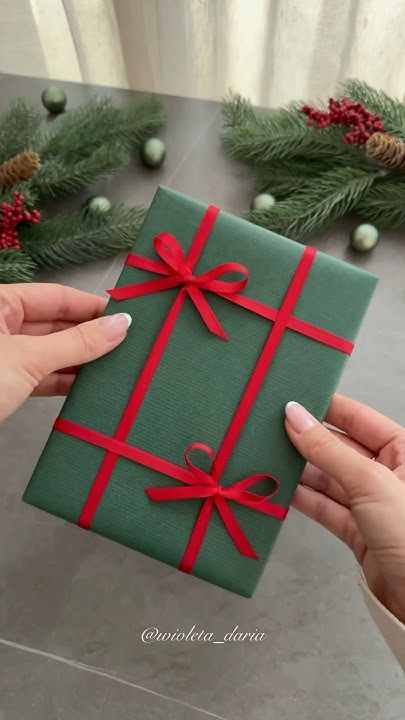 two hands holding a green wrapped gift box with red ribbon and bow on it, surrounded by evergreen branches